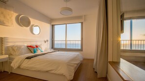 Un dormitorio con una cama grande, una mesita de noche, una ventana con vista al mar y un balcón con barandilla.