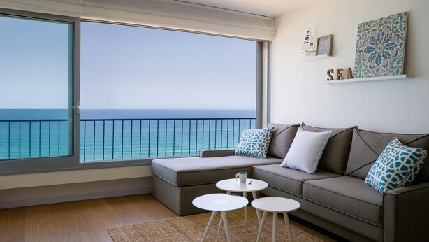 Una sala de estar moderna con un ventanal que da al mar, un sofá gris con cojines estampados y una pequeña mesa blanca.