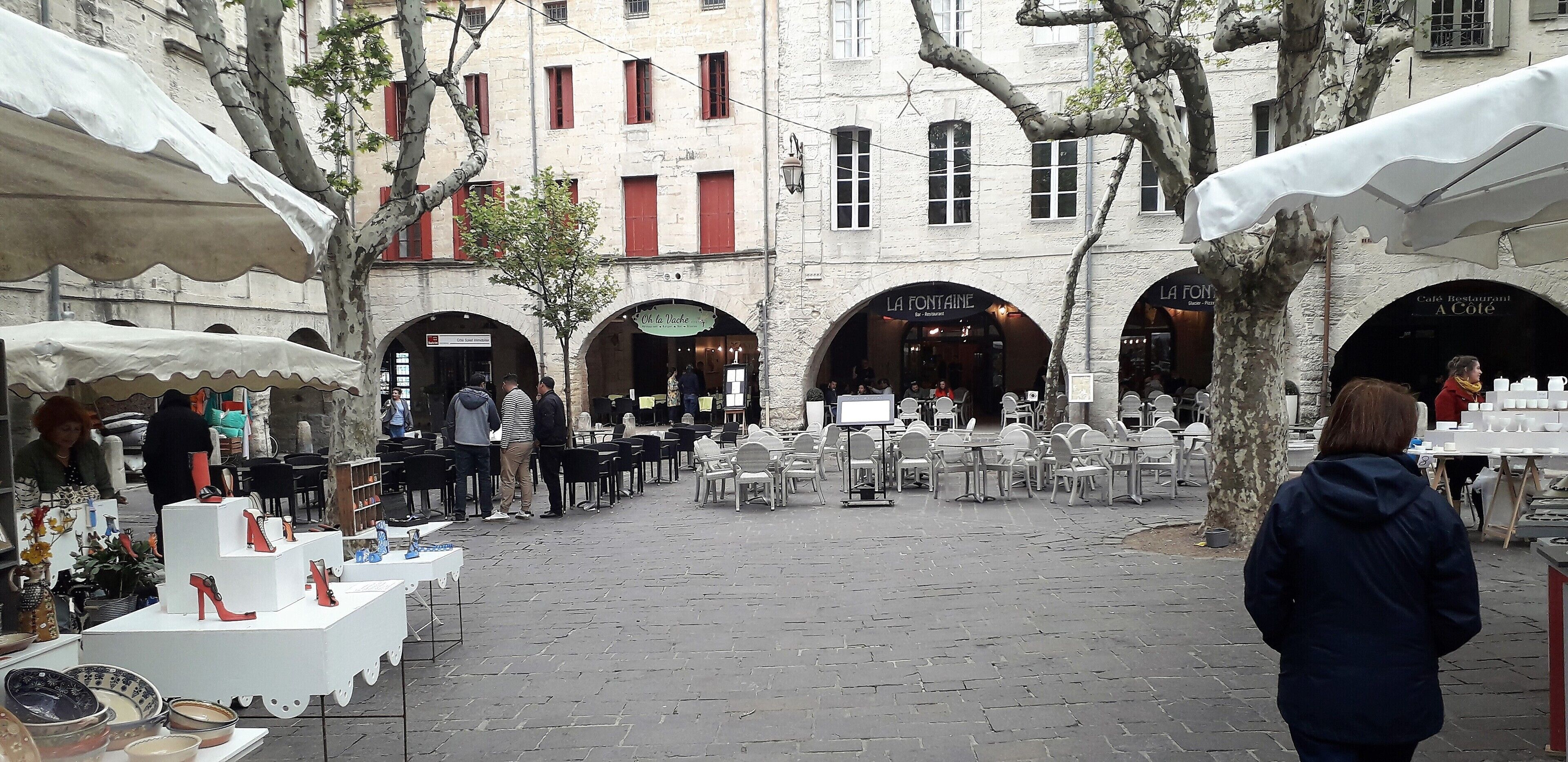 An outdoor market with tables displaying shoes and other items, surrounded by buildings with arches and a cafe named "La Fontaine".