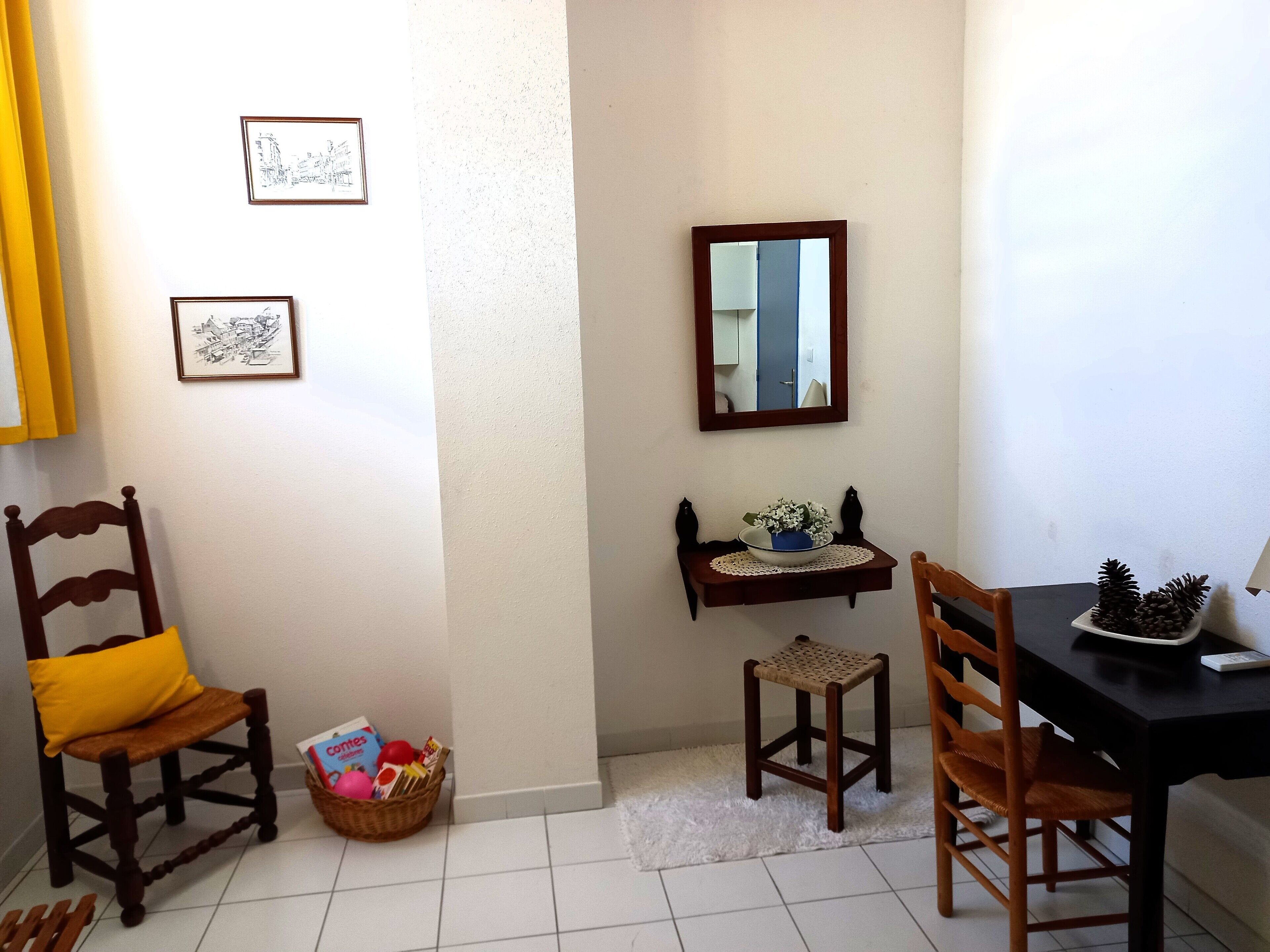 A dining area with a table, chairs, a small shelf with decorative items, and framed pictures on the wall.