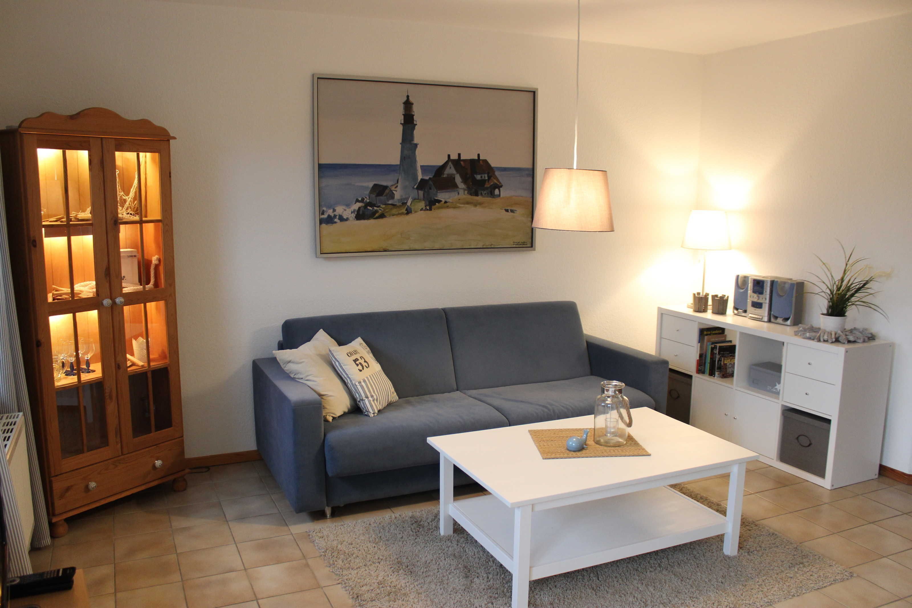A living room with a blue sofa, a white coffee table, a wooden cabinet with glass doors, a painting of a lighthouse, and a lamp.