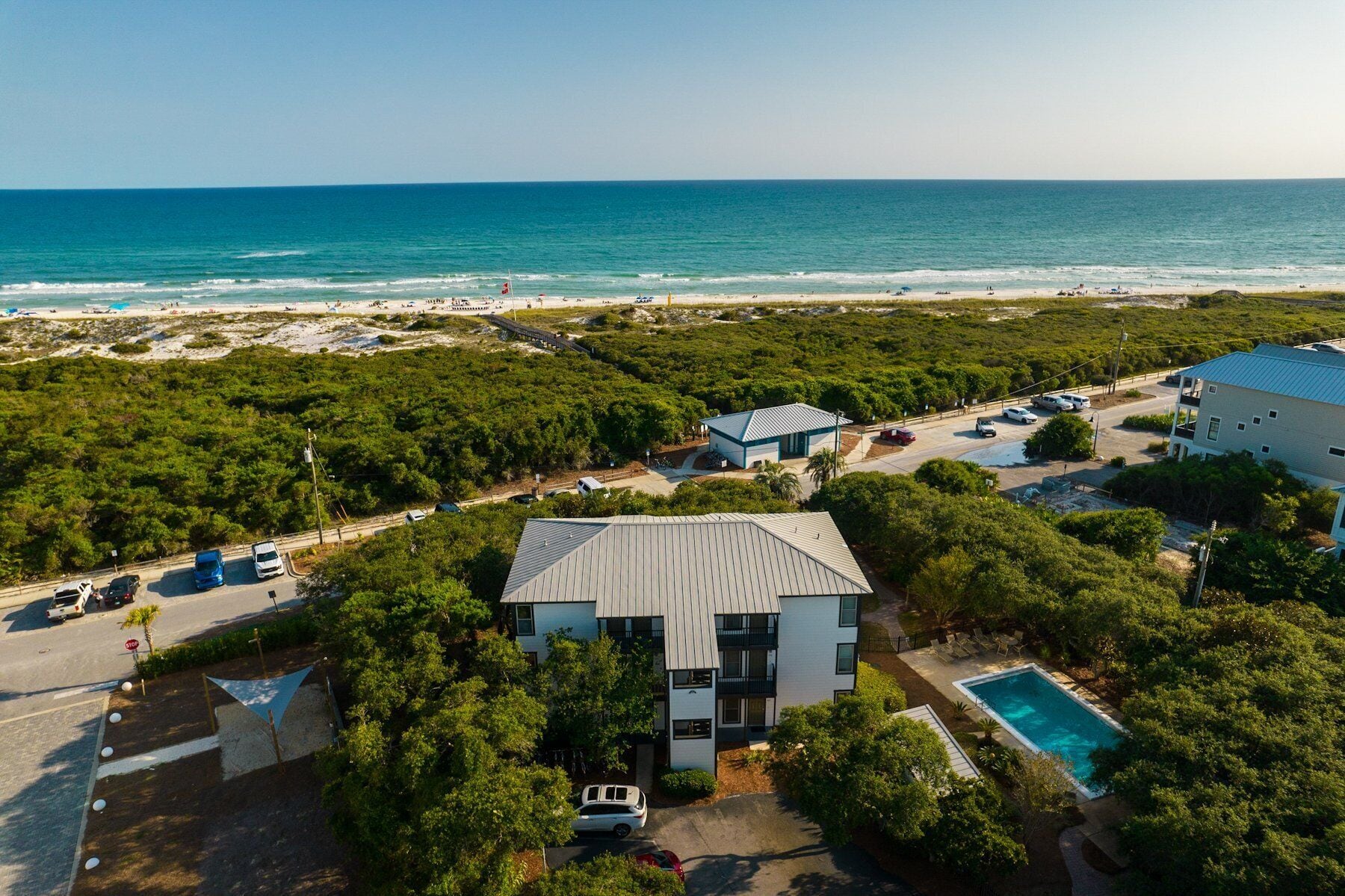 A residential area with a large house featuring a swimming pool, surrounded by lush greenery and a view of the beach and ocean.
