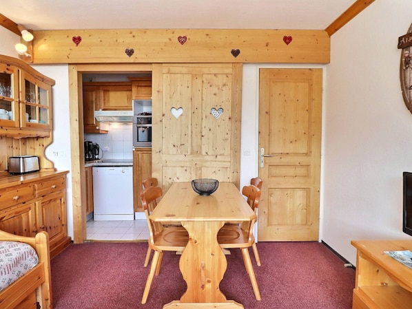 A wooden dining table with chairs, a kitchen area with cabinets and appliances, and a red carpeted floor.
