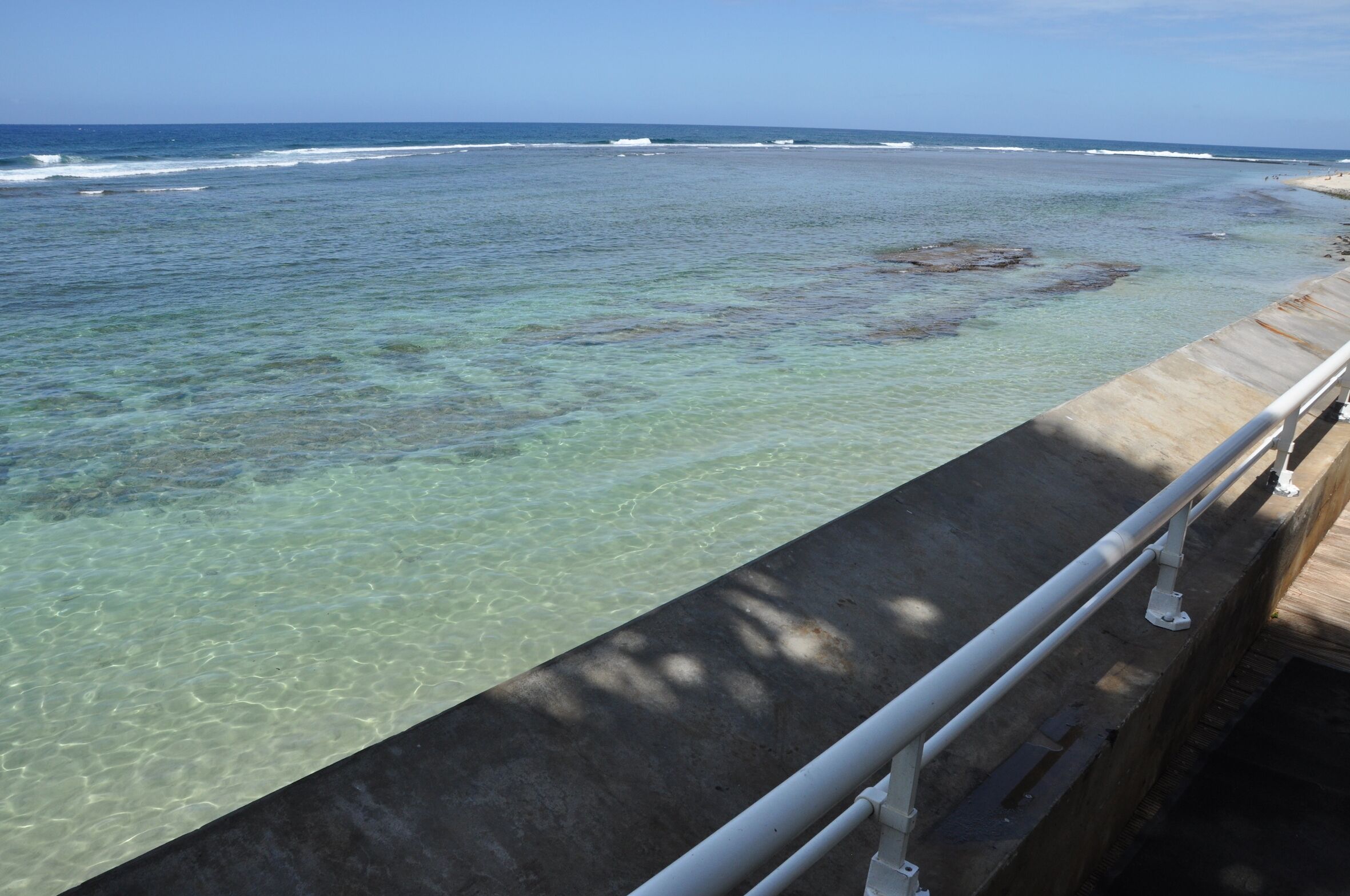 A coastal view with clear water, a concrete barrier, and a metal railing.