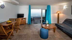A living room with a TV, wooden furniture, a sofa, a table with a vase, and a view of the ocean through a glass door. - REVE AUSTRAL T2 C au bord du Lagon 180° sea view (St Pierre)