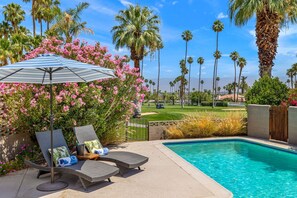 Zona junto a la piscina con sillones de descanso, una sombrilla a rayas y vistas a un campo de golf.