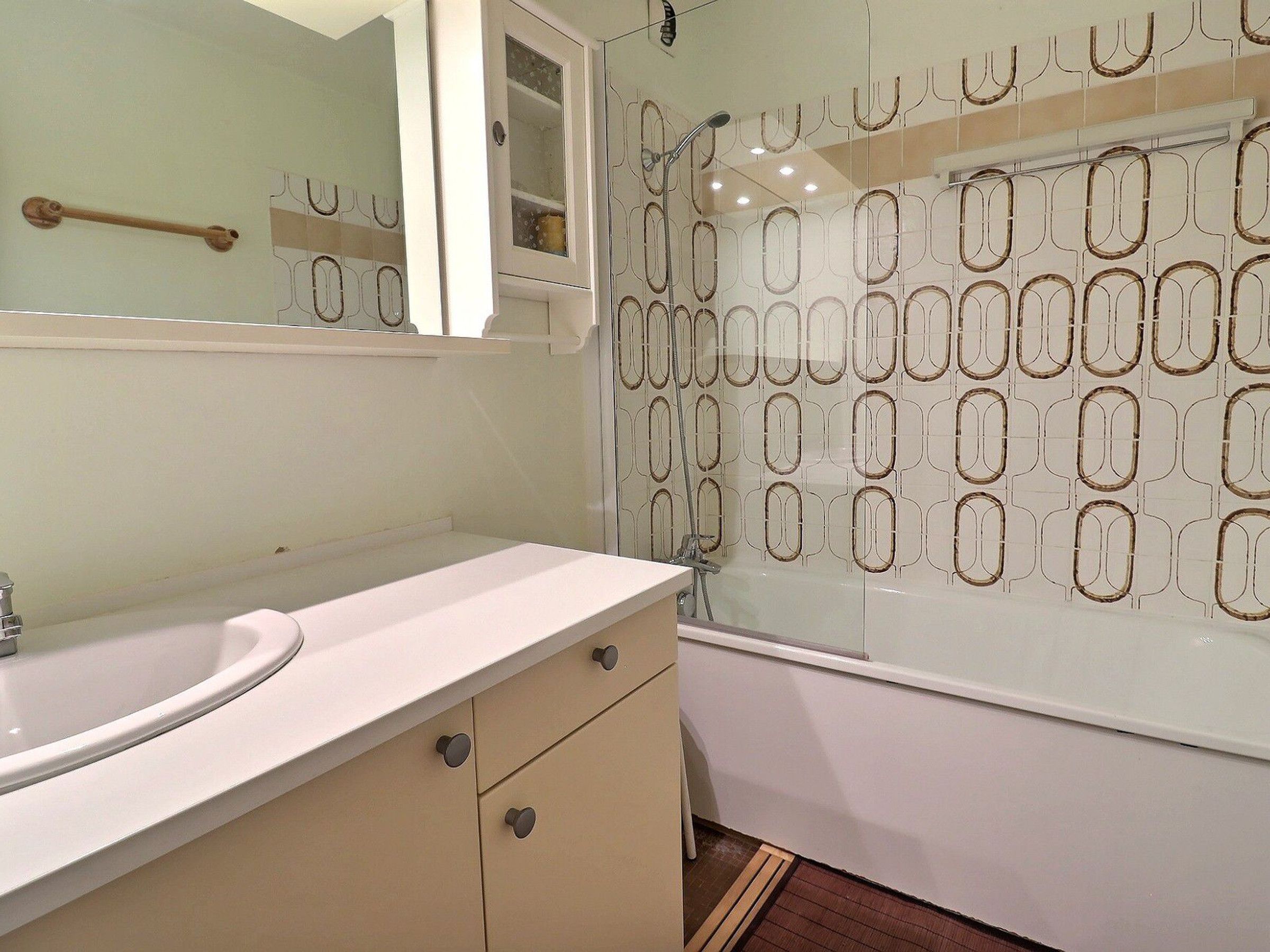A bathroom with a white sink, beige cabinets, a mirror, and a shower with a patterned glass door.