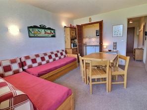 A cozy room with a red and white checkered sofa, a wooden dining table with chairs, a kitchenette with a refrigerator and stove, and a wall-mounted clock.