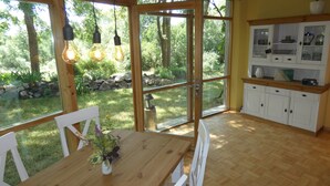 Une table à manger en bois avec des chaises, un buffet blanc aux portes vitrées et une vue sur un jardin par de grandes fenêtres.
