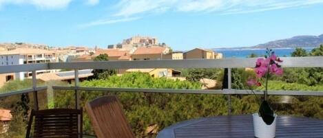Un balcón con una mesa de madera redonda y dos sillas, con vista a un pueblo costero con edificios y vegetación.