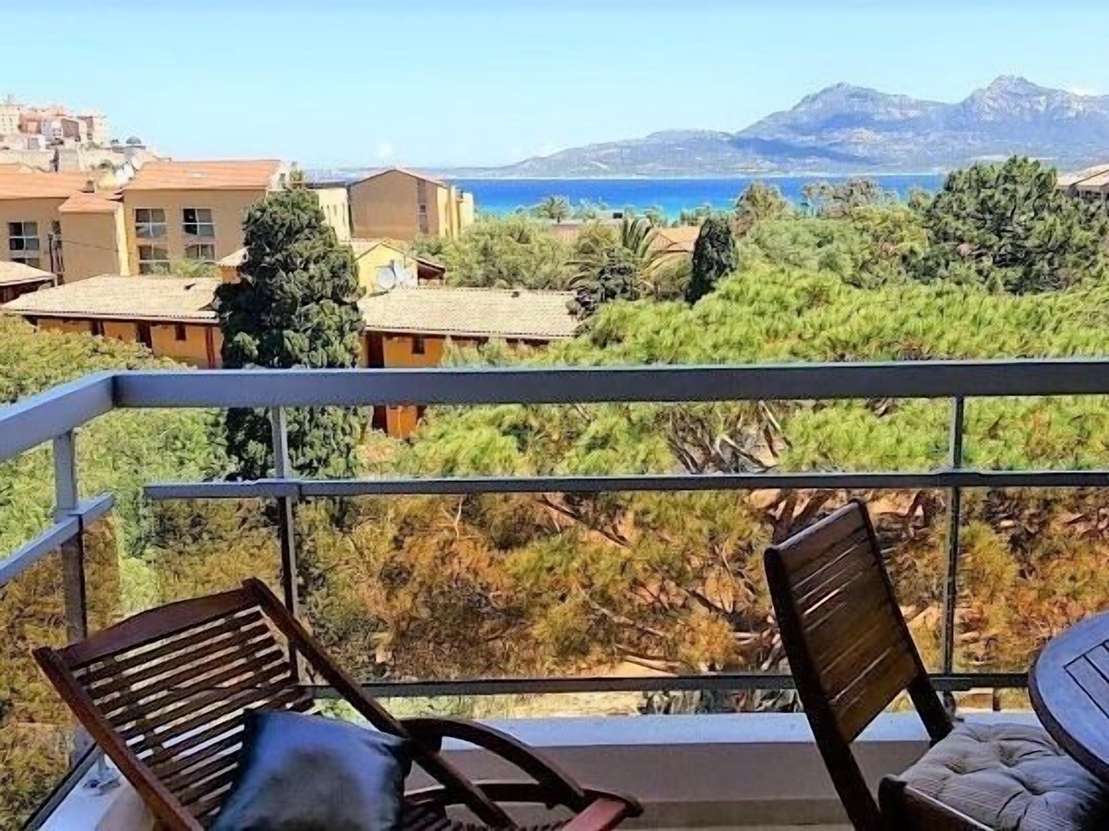 A balcony with two wooden chairs, overlooking a coastal landscape with buildings and trees.