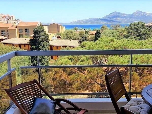 A balcony with two wooden chairs, overlooking a coastal landscape with buildings and trees.