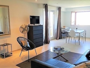 Un salon moderne comprenant un canapé, une table basse, un téléviseur posé sur un meuble, un coin repas avec des chaises et une grande fenêtre.
