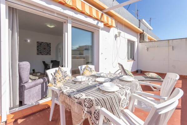 An outdoor patio with a table set for dining, surrounded by white chairs, and a sliding glass door leading to an interior space.