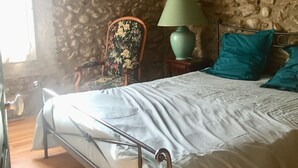 Una camera da letto con parete in pietra, pavimento in legno, struttura del letto in metallo, lampada verde e poltrona con motivi floreali.
