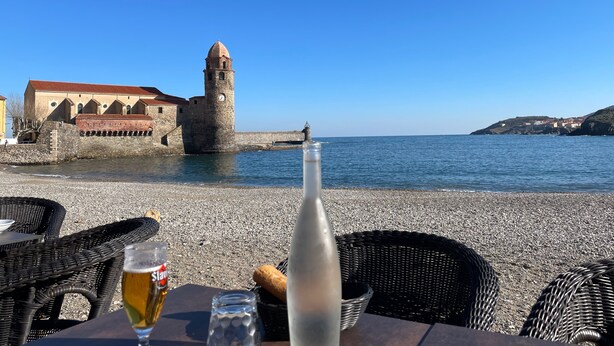 Vista sul mare con una torre storica, un tavolo con una bottiglia e un bicchiere, e sedie in vimini.