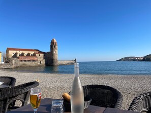 A seaside view with a historic tower, a table with a bottle and a glass, and wicker chairs.