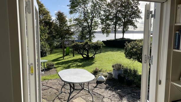 Une terrasse avec une table et des chaises en pierre, un petit jardin avec une aire de jeux et une vue sur un lac.