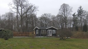 Une petite maison à deux étages, dotée d’un toit en pente, entourée d’une cour verdoyante et de grands arbres.