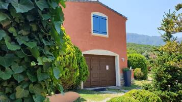 Une maison à deux étages avec une porte de garage marron, un volet bleu et un jardin bien entretenu, agrémenté de divers arbustes et d’un grand arbre.