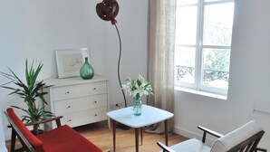 Una habitación con piso de madera, una mesa blanca, dos sillas, un armario blanco pequeño, un jarrón verde y una ventana con cortinas.