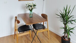 Un comedor con una mesa de madera, dos sillas negras, un florero con flores, una planta en maceta, un espejo y un cuadro en la pared.