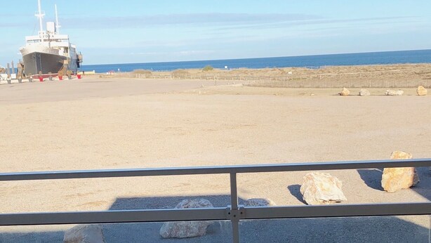 A beach with a ship in the background, a railing in the foreground, and a clear sky.