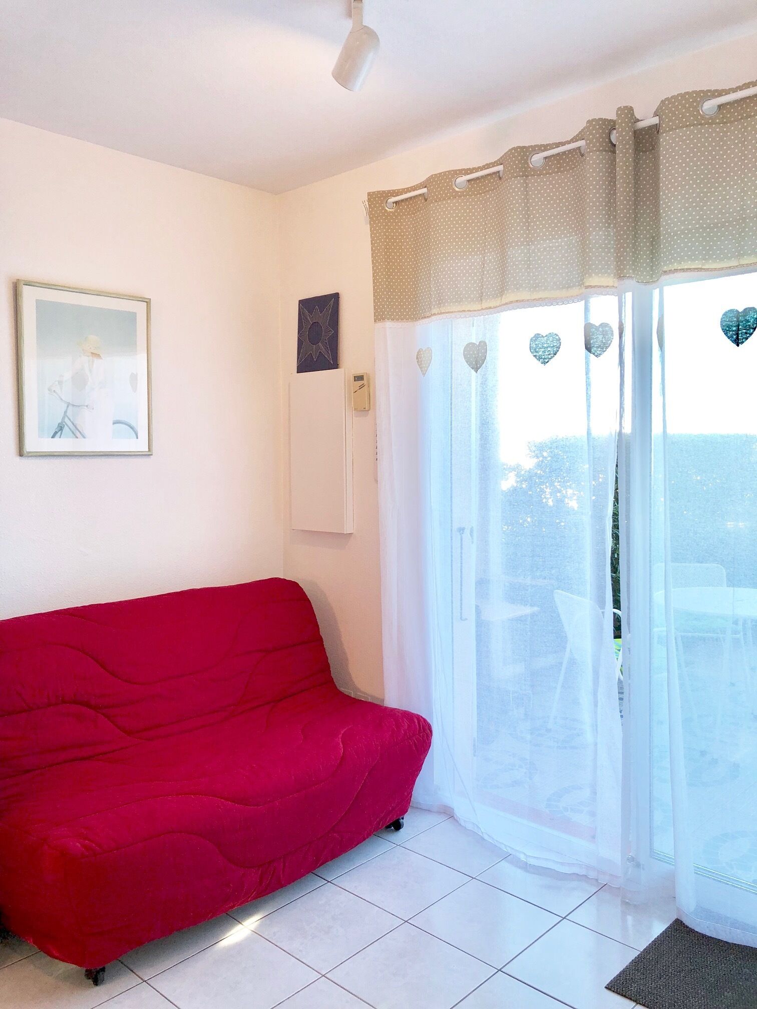 Ein Zimmer mit einem roten Sofa, einem Deckenventilator, einem gerahmten Bild an der Wand und einem Fenster mit herzförmigen Verzierungen.