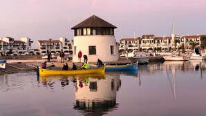 A marina with boats docked, a small building with a conical roof, and a row of residential buildings in the background.