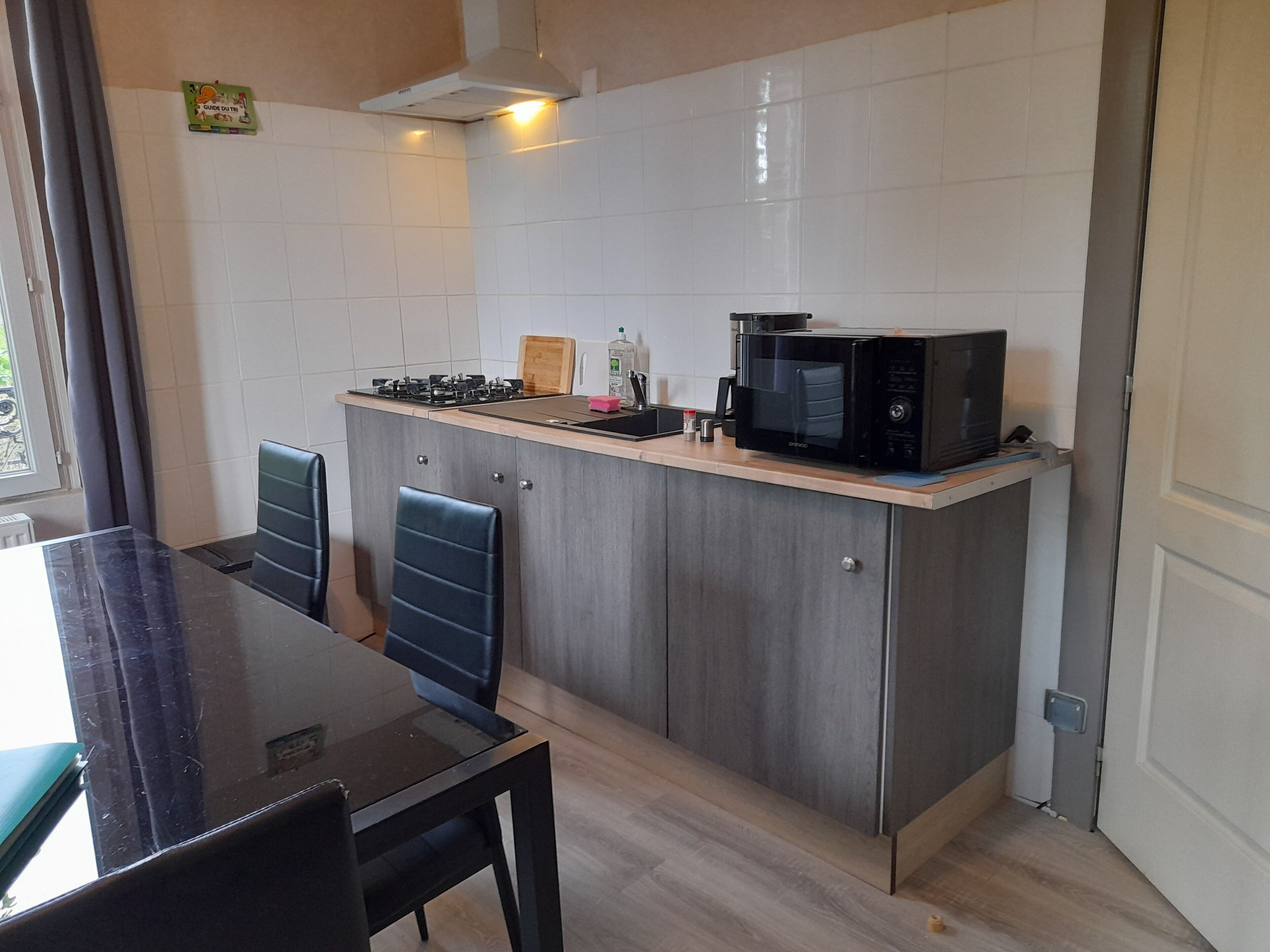 Una cocina compacta con estufa a gas, microondas y una pequeña encimera. Hay una mesa de comedor con sillas y una ventana con cortinas.
