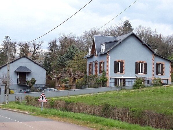 Een woonwijk met twee huizen, een met een grijze gevel en de andere met een blauwe gevel en rode luiken. Er staat een driehoekig waarschuwingsbord bij de weg.