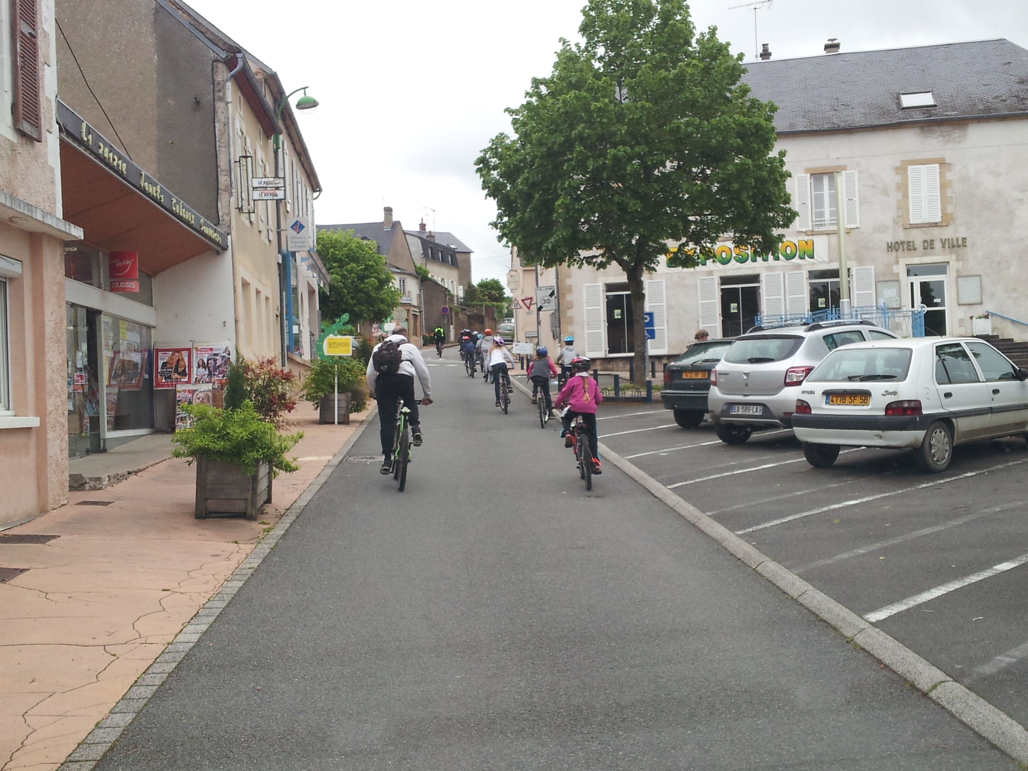 A street with cyclists, parked cars, and buildings including a hotel.