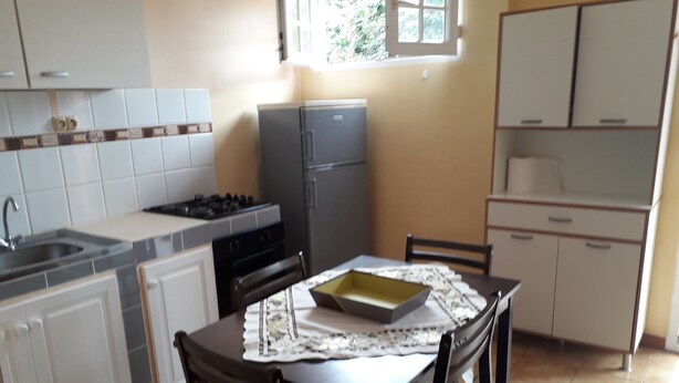 Une cuisine compacte équipée d’une cuisinière à gaz, d’un évier et d’un réfrigérateur. On y trouve une table à manger avec des chaises, et une fenêtre située au-dessus de l’évier.