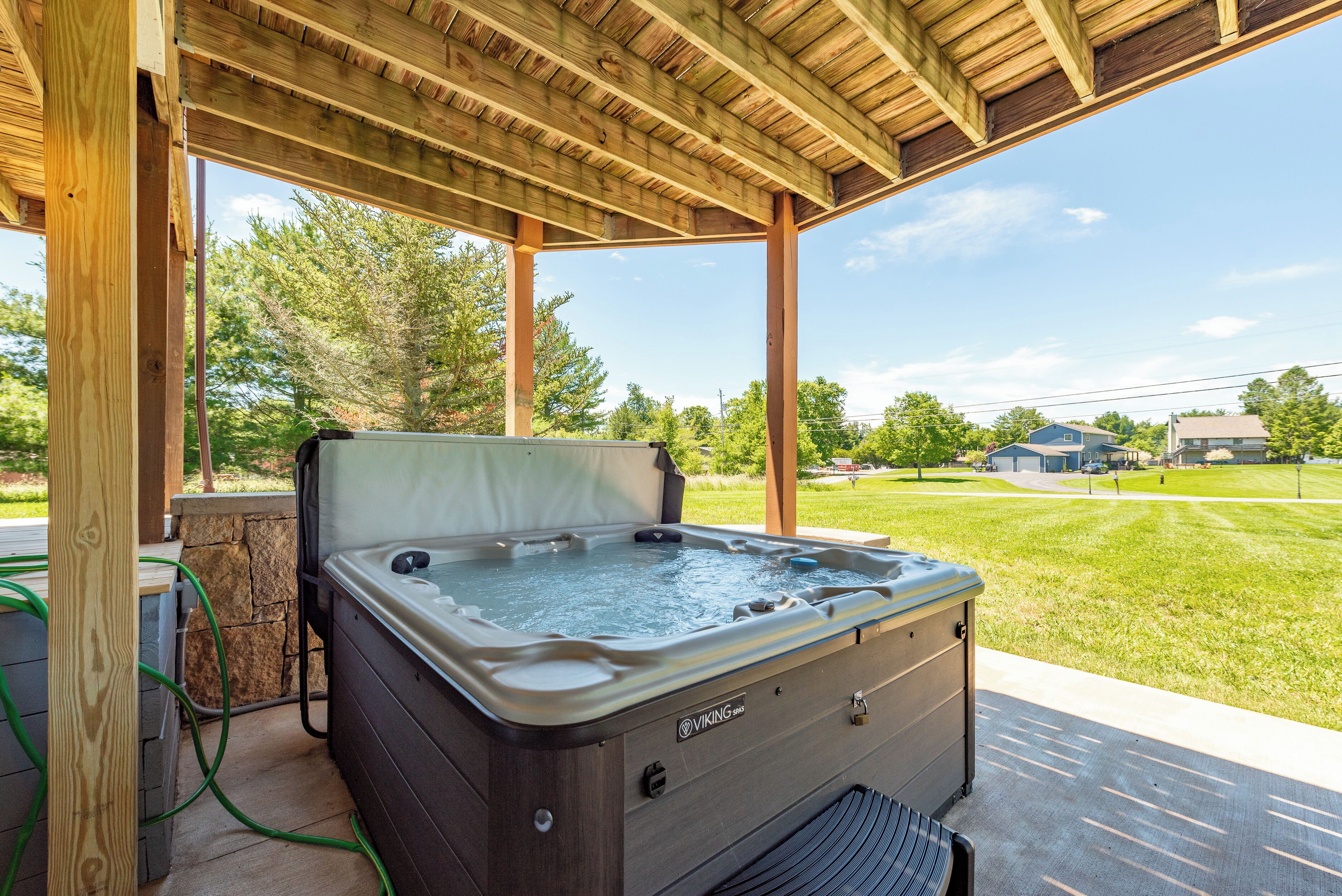 A hot tub under a wooden pergola with a stone wall and a grassy yard in the background.