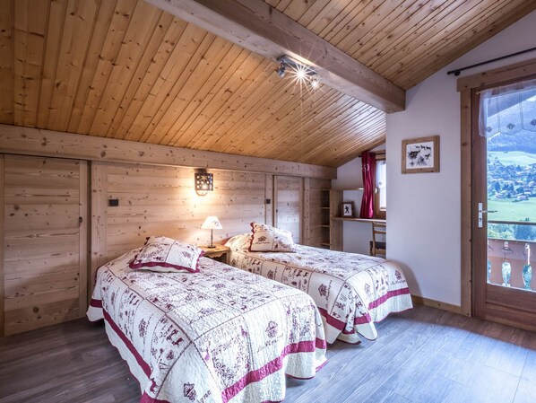 A cozy bedroom with two beds, wooden ceiling beams, a nightstand with a lamp, a small desk, and a window with a view of the countryside. - Chalet 4 bdr, near slopes, family/friends ideal, WIFI (La Clusaz)