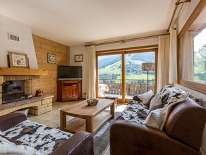 A cozy living room with a fireplace, a television, and a scenic view through the glass doors. - Chalet 4 bdr, near slopes, family/friends ideal, WIFI (La Clusaz)