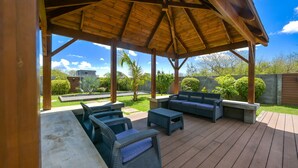 Un patio techado al aire libre con mobiliario de mimbre y una zona de asientos de concreto, con vista a un jardín ajardinado.