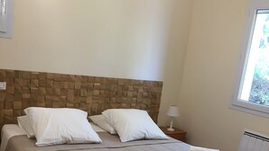 A neatly made bed with white linens and towels, a wooden headboard, and a window with a view of greenery.