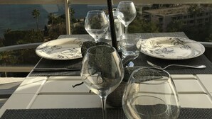 A table set for two with white plates, clear wine glasses, and a vase with white flowers. A view of the ocean and palm trees is visible through a balcony railing. - SUPER CANNES / GOLFE JUAN LUXURY APARTMENT SEA IMPRENABLE SEA. PRIVATE FLOOR (Vallauris)