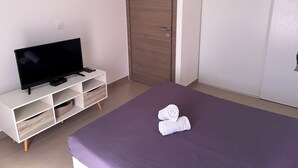 Une chambre à coucher avec un lit, un téléviseur posé sur un meuble blanc, une armoire en bois et une couvre-lit violette.