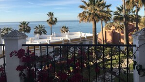 A balcony with a view of the sea, palm trees, and a white building with yellow accents.