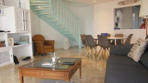 A modern living room with a glass coffee table, a sofa, a dining area with chairs, a staircase, and a bookshelf.