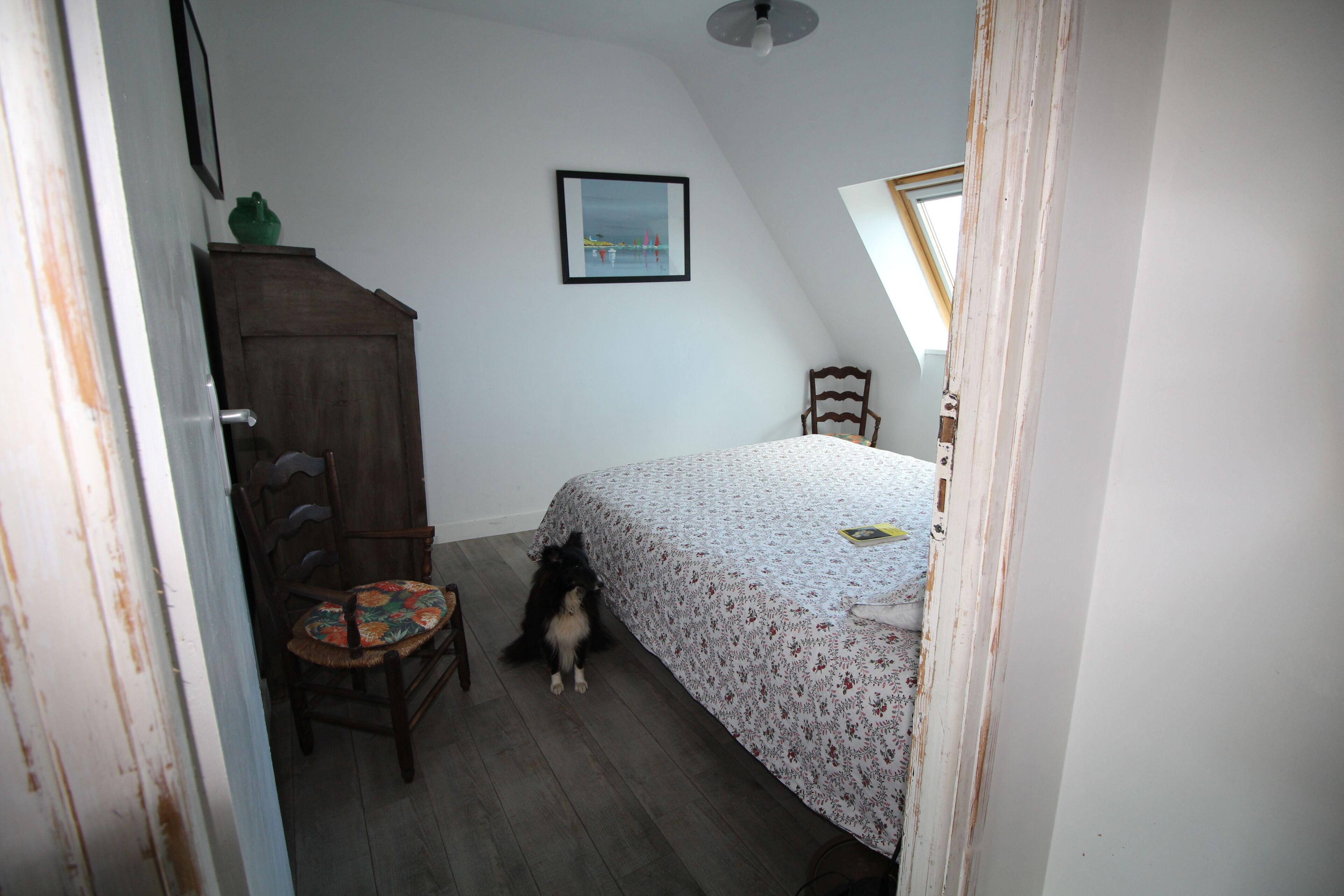 A bedroom with a bed, a chair, a wooden cabinet, a framed picture on the wall, and a skylight.