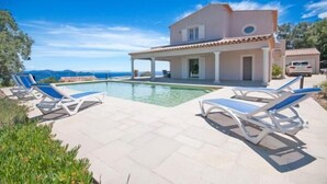 A two-story house with a swimming pool, lounge chairs, and a view of the sea.