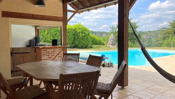 An outdoor patio with a wooden table and chairs, a barbecue grill, and a view of a swimming pool and greenery. - Cottage rental between Cahors and St Cirq Lapopie (Arcambal)