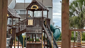 Una estructura de juego de madera con tobogán y elementos para escalar, con un entorno residencial al fondo.