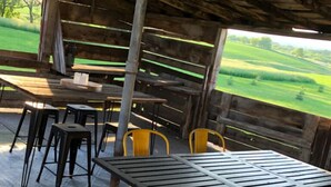 Eine Terrasse im Freien mit Tisch und Stühlen, Blick auf ein grünes Feld und eine Holzhütte.
