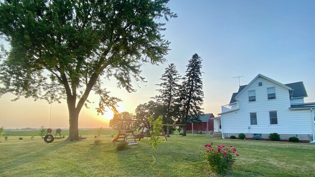 Ein grasbewachsenes Feld mit einer Schaukel, einem großen Baum und einem weißen Haus mit einem roten Bauernhof im Hintergrund.