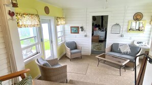 Ein gemütliches Wohnzimmer mit Korbmöbeln, einer Couch und einem Sessel. Ein Fenster mit Vorhängen, eine Tür und ein kleiner Tisch sind ebenfalls vorhanden.