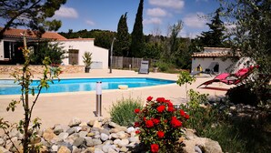 A swimming pool surrounded by rocks and flowers, with a white building and a red chair in the background. - Quiet cottage + pool, for 3 pers. (Lézignan-Corbières)